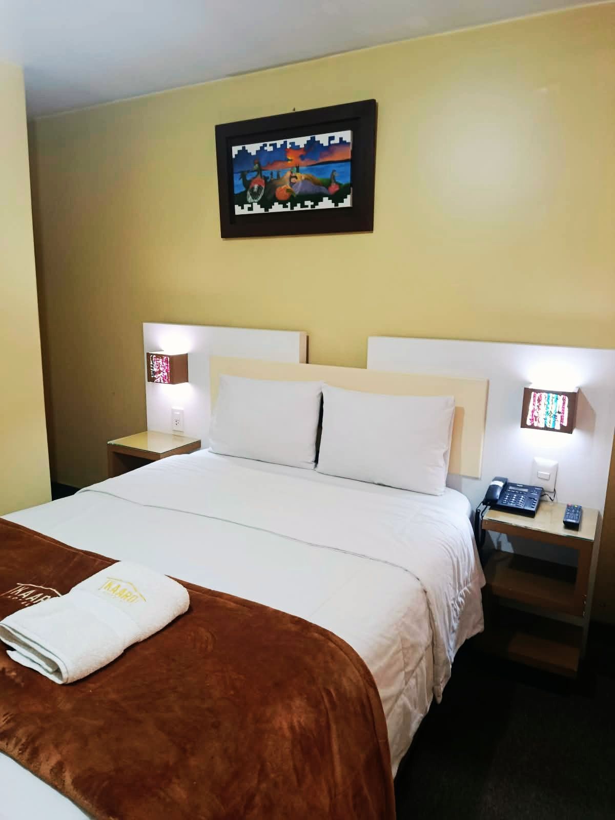 A hotel room with a bed, two bedside tables with lamps, a painting on the wall, and a folded towel on the bed.