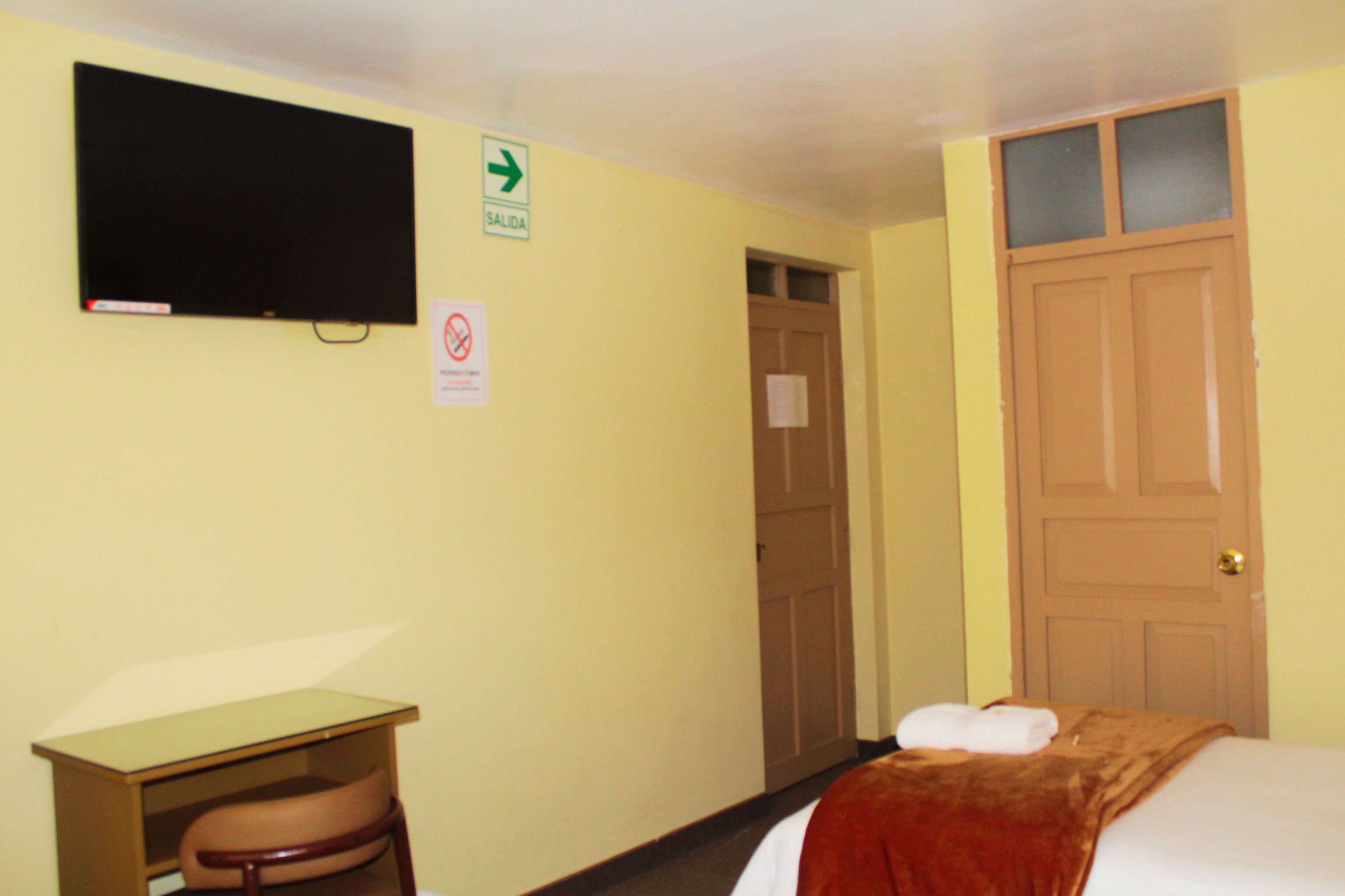 A hotel room with a bed, a desk, a television mounted on the wall, and a door with a sign indicating the exit.