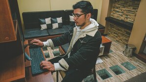 En person sidder ved et skrivebord med en computer i et rum med en sofa og en stenspir.