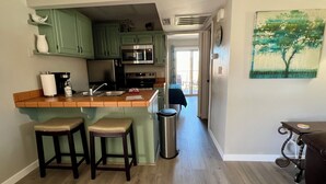 Eine Küche mit grünen Schränken, einem Edelstahlkühlschrank und einer Holz Arbeitsplatte. Zwei Hocker und ein Mülleimer sind ebenfalls vorhanden.