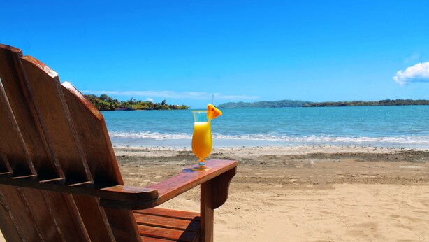 Una silla de playa de madera con un vaso de jugo de naranja y una pajita en una playa arenosa bajo un cielo azul despejado y un mar tranquilo.