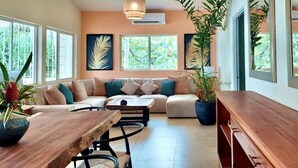 Una sala moderna con un sofá, una mesa de centro y una mesa de comedor. Hay ventanas grandes, una lámpara colgante y plantas en macetas.