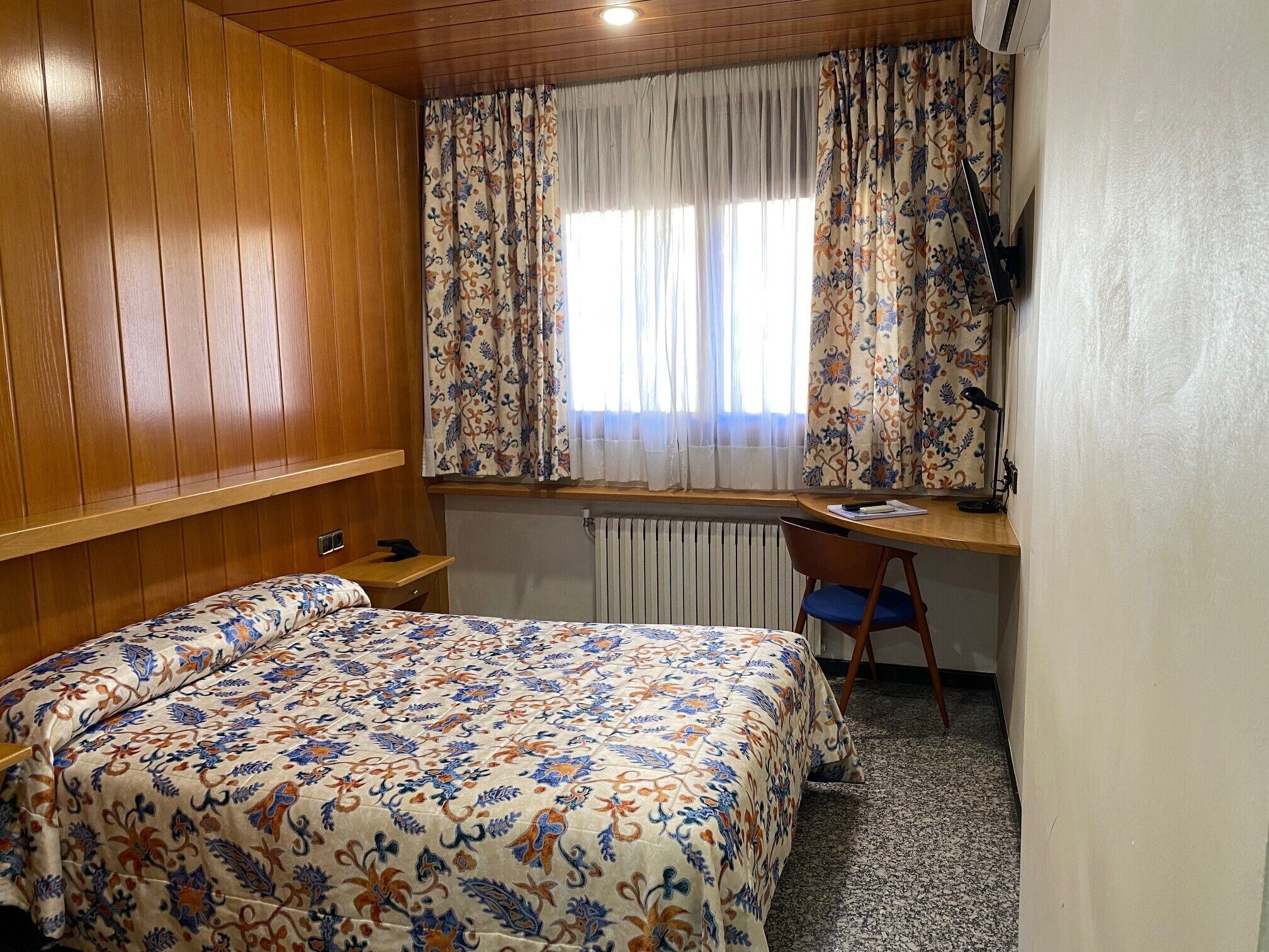 A bedroom with a wooden headboard, a bed with a floral bedspread, a desk with a chair, and a radiator under a window with patterned curtains.
