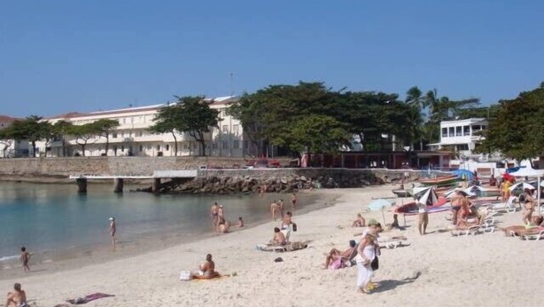 Een strand met mensen die zonnen en zwemmen, een stenen muur en gebouwen op de achtergrond.