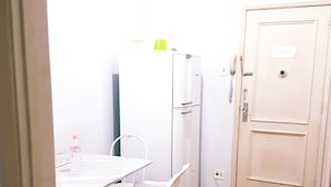 Een kleine, witte kamer met een betegelde vloer, een witte tafel met twee stoelen, een witte koelkast en een deur met een bordje.