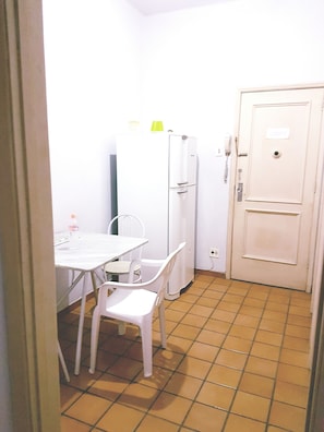 Een kleine, witte kamer met een betegelde vloer, een witte tafel met twee stoelen, een witte koelkast en een deur met een bordje.