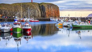A harbor with boats docked, a lighthouse, and a cliff in the background.