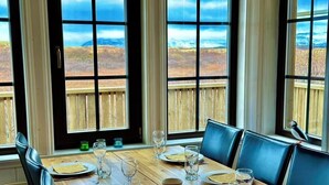 A dining table set for six with plates, glasses, and bowls, overlooking a scenic landscape through large windows.