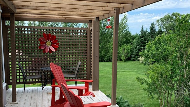 Un espacio al aire libre techado con sillas Adirondack rojas y una mesa, rodeado por una terraza de madera y un cerramiento de celosía con una decoración floral roja.