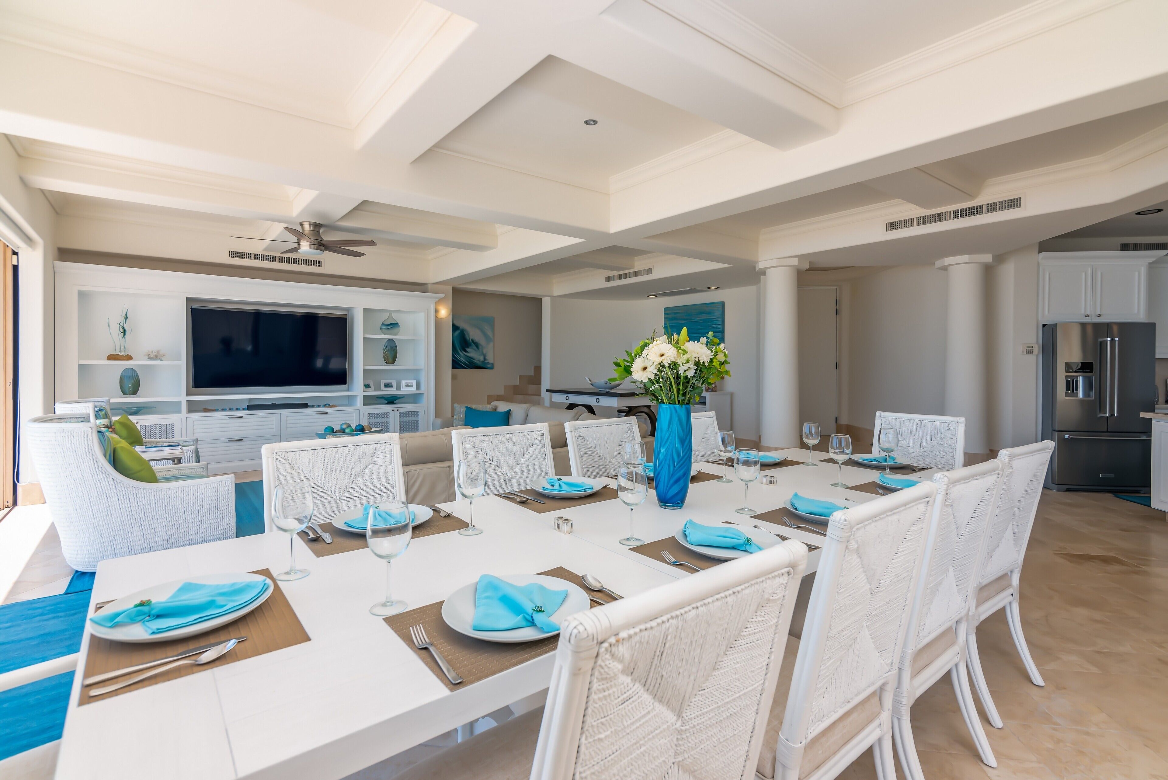 Un coin repas avec une grande table dressée pour six personnes, des chaises en osier blanches, un vase bleu rempli de fleurs et, au fond, une cuisine moderne.
