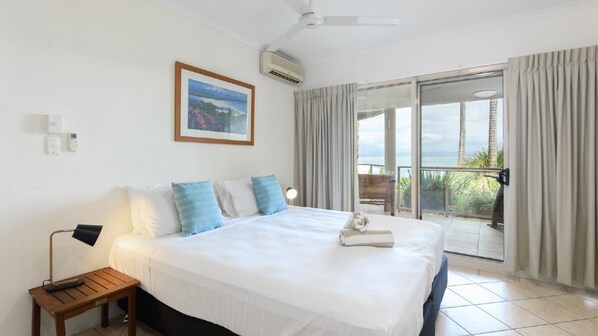Une chambre d’hôtel avec un grand lit, une table de chevet, un ventilateur de plafond, un appareil de climatisation, un tableau encadré au mur et un balcon avec des rideaux.