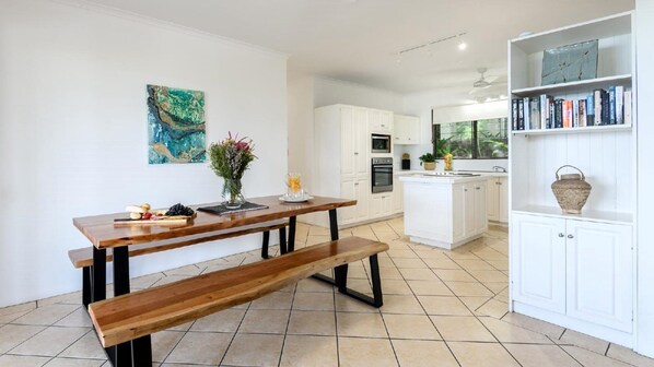 Una cocina moderna con armarios blancos, una mesa de comedor de madera con bancos, una pequeña isla con fregadero y una estantería con libros y objetos decorativos.