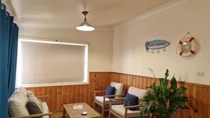 A room with a wooden paneled wall, a ceiling fan, a large window with curtains, a sofa, two armchairs, a small table, and a potted plant. - Seahaven Beach House - This brilliant beach house sleeps up to 8 adults and 2 children. (shellharbour)