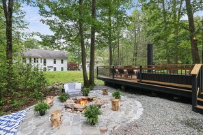 Un foyer en pierre entouré de plantes, une terrasse en bois avec des sièges et une maison en arrière-plan.