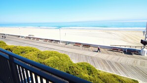 Vista a la playa desde un balcón con terraza de madera, bancos y cielo despejado.