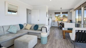 A modern living room with a grey sofa, a small ottoman, a dining table with chairs, and a view of the outdoors through large windows. - Serindipity - 180 Degrees ocean view (Yanchep)