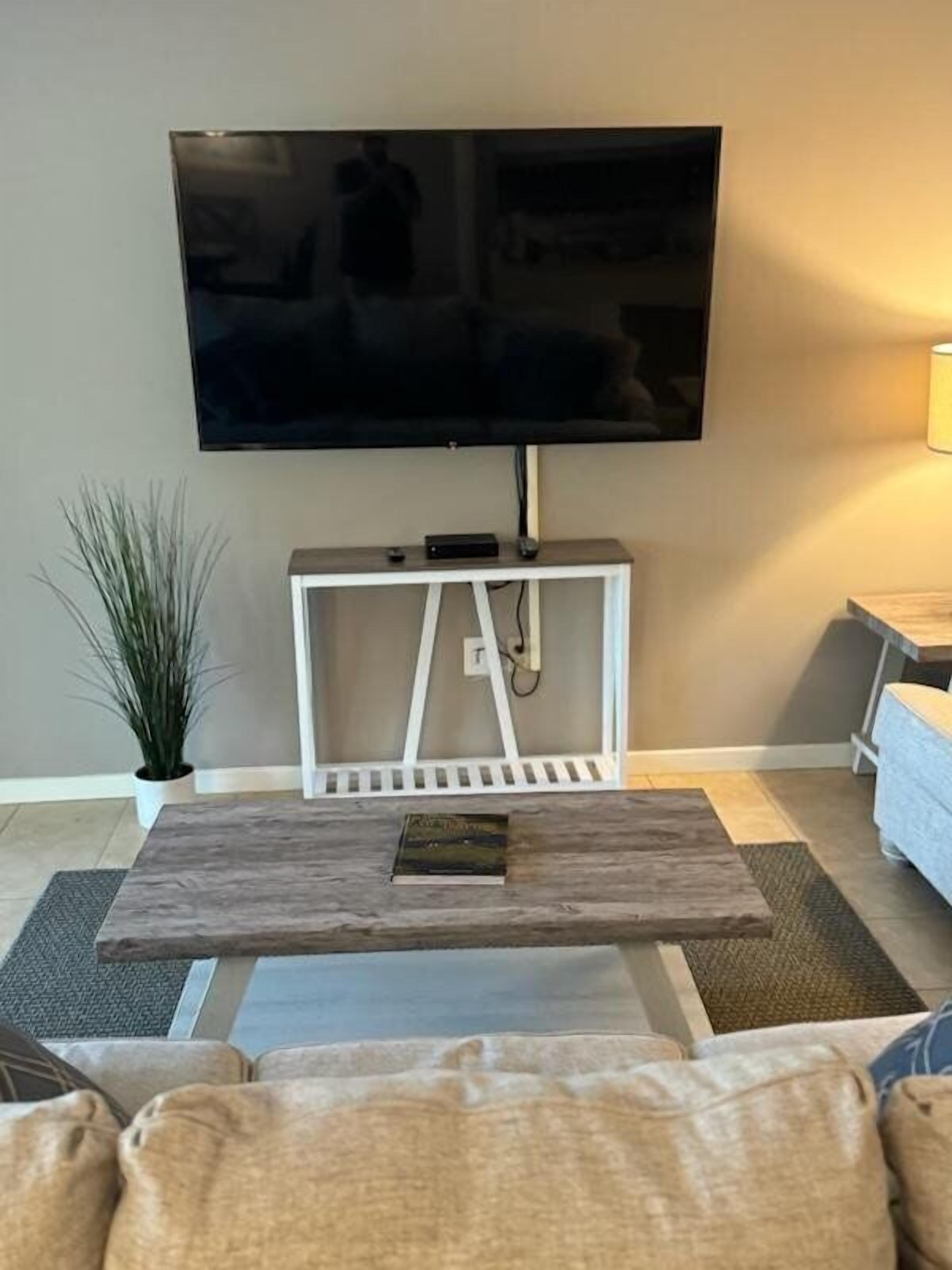 A living room with a flat-screen TV mounted on a white stand, a coffee table with a book, and a potted plant.