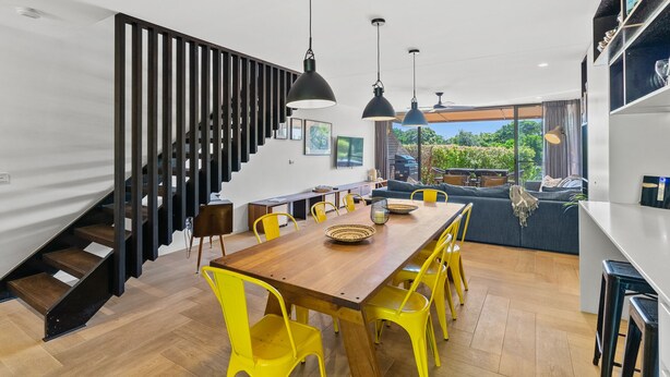 A modern kitchen with a wooden dining table, yellow chairs, and a staircase with black railings. - Surfside 6 at James Cook - Clarkes Beach (Byron Shire)