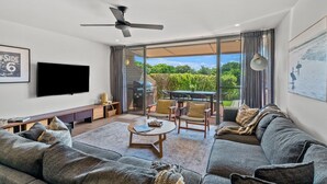 A modern living room with a flat-screen TV, a ceiling fan, and a large window overlooking a garden. - Surfside 6 at James Cook - Clarkes Beach (Byron Shire)