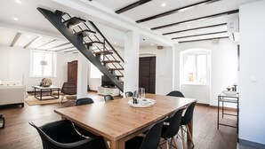 Amplia sala de estar con una mesa de comedor de madera, sillas negras, una escalera y una cocina con encimera y lavamanos blancos.