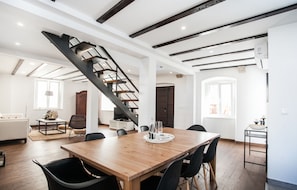 Een ruime woonkamer met een houten eettafel, zwarte stoelen, een trap en een keuken met een wit aanrecht en gootsteen.