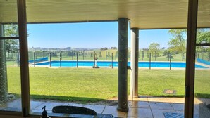 Vista desde una ventana sobre una piscina, un área con césped y colinas al fondo.