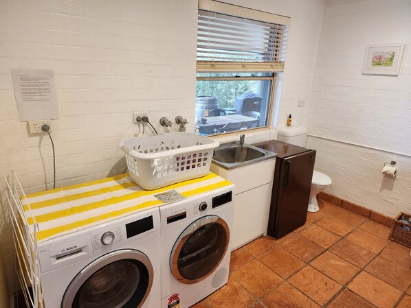 A compact laundry room with a Bosch washing machine, a dryer, a small sink, and a window with blinds.
