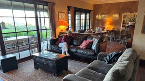 A spacious living room with a large window, leather sofas, a coffee table, and a dining area with a chandelier. - Glyn Newydd - is the perfect place to unwind with family & friends and pets (Megalong Valley)