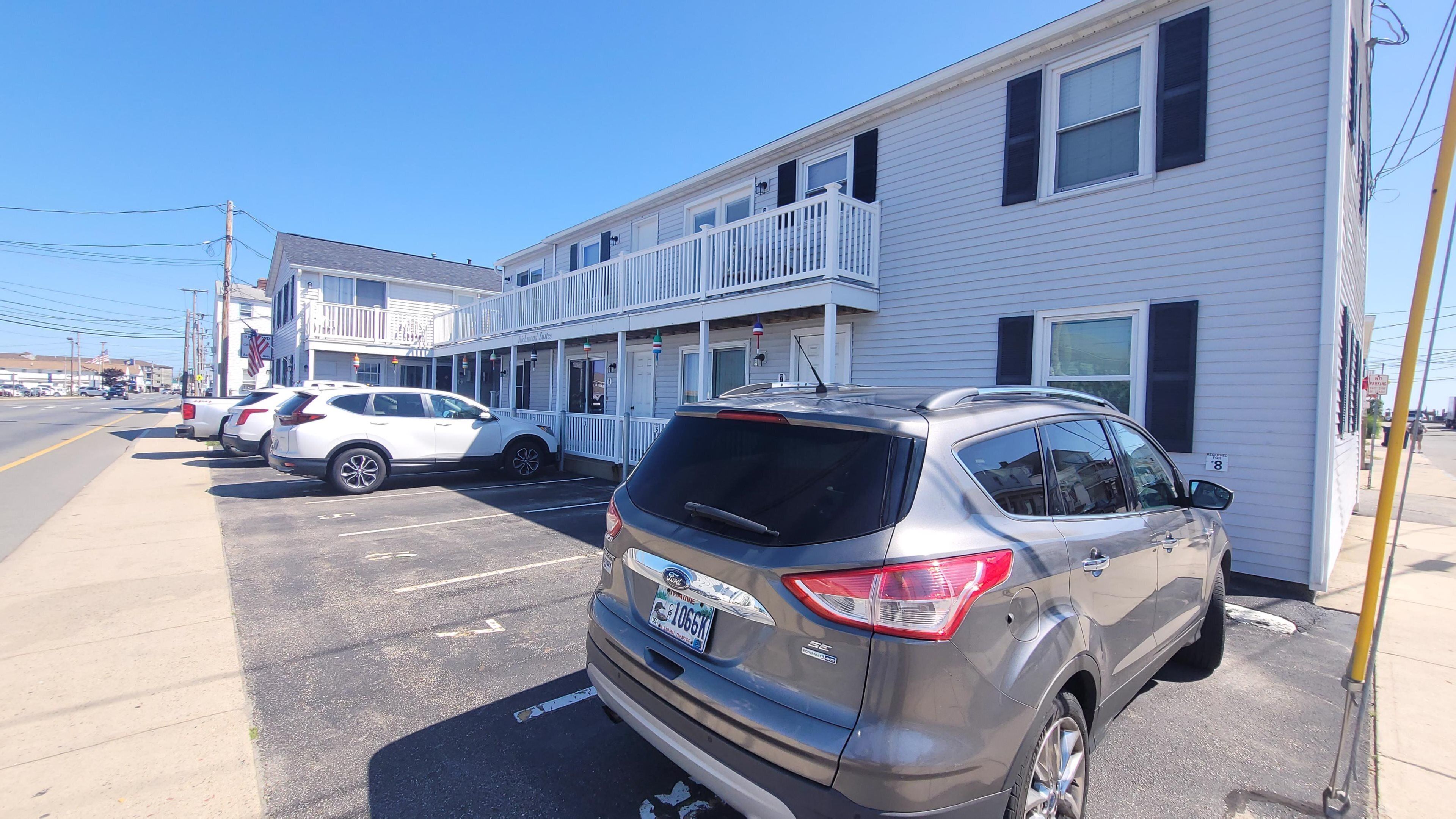 A parking lot with several cars, a building with a white facade and a balcony, and a clear blue sky.