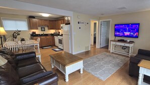 Ein Wohnzimmer mit einer Couch, einem Couchtisch und einem Fernseher. Im Hintergrund ist eine Küche mit Holzschränken und einem Kühlschrank zu sehen.