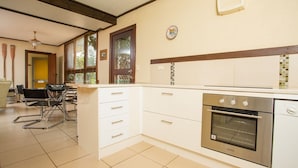 A modern kitchen with white cabinets, a built-in oven, and a dining area with a table and chairs. - Banksia Seven - Merimbula (Merimbula)