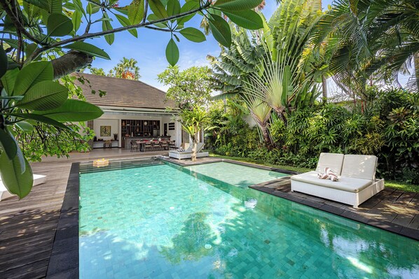 A swimming pool with a wooden deck, a lounge area with a sofa and chairs, and a dining area with a table and chairs.