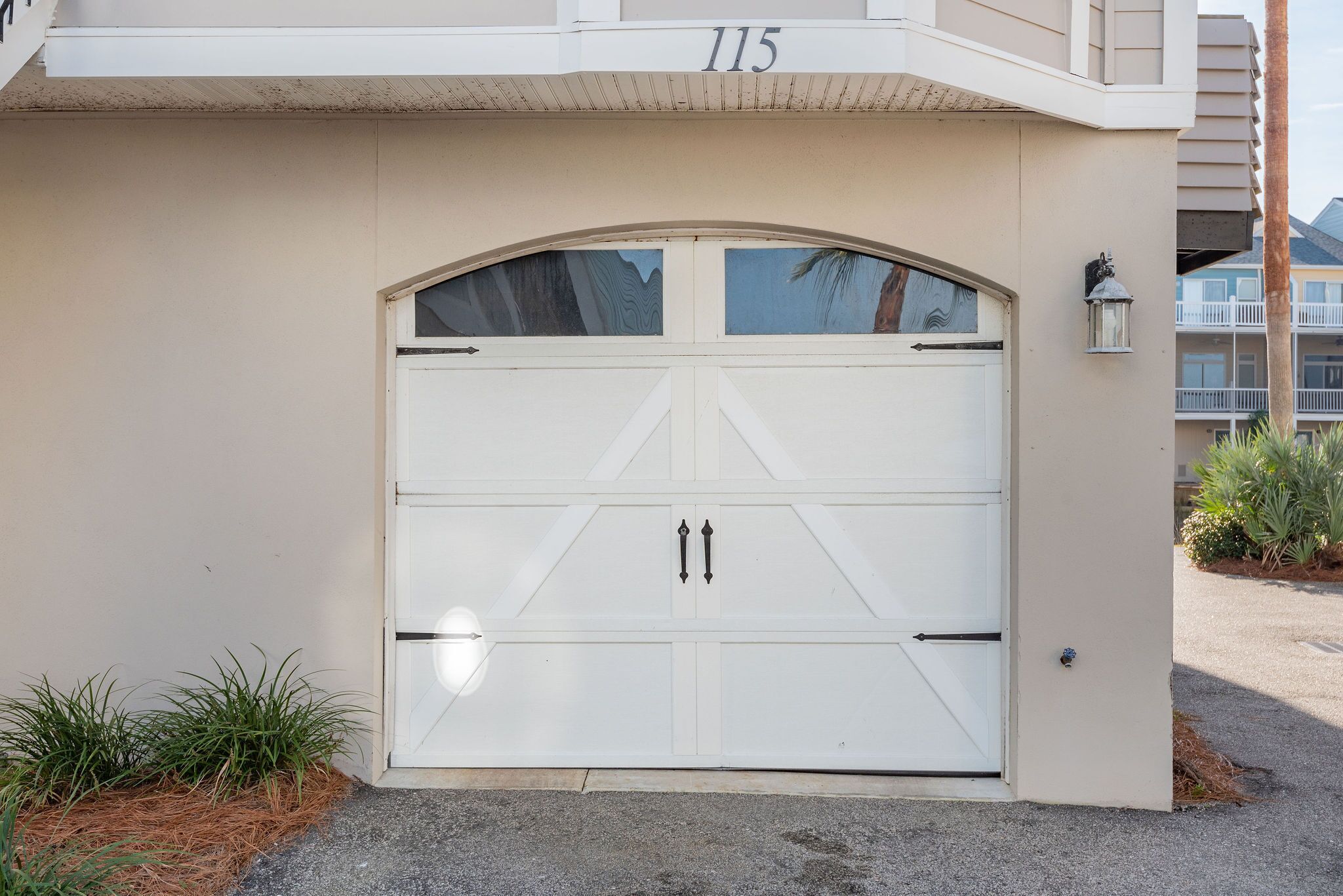 A white garage door with a number "115" above it, a small planter with plants beside it, and a building with balconies in the background.