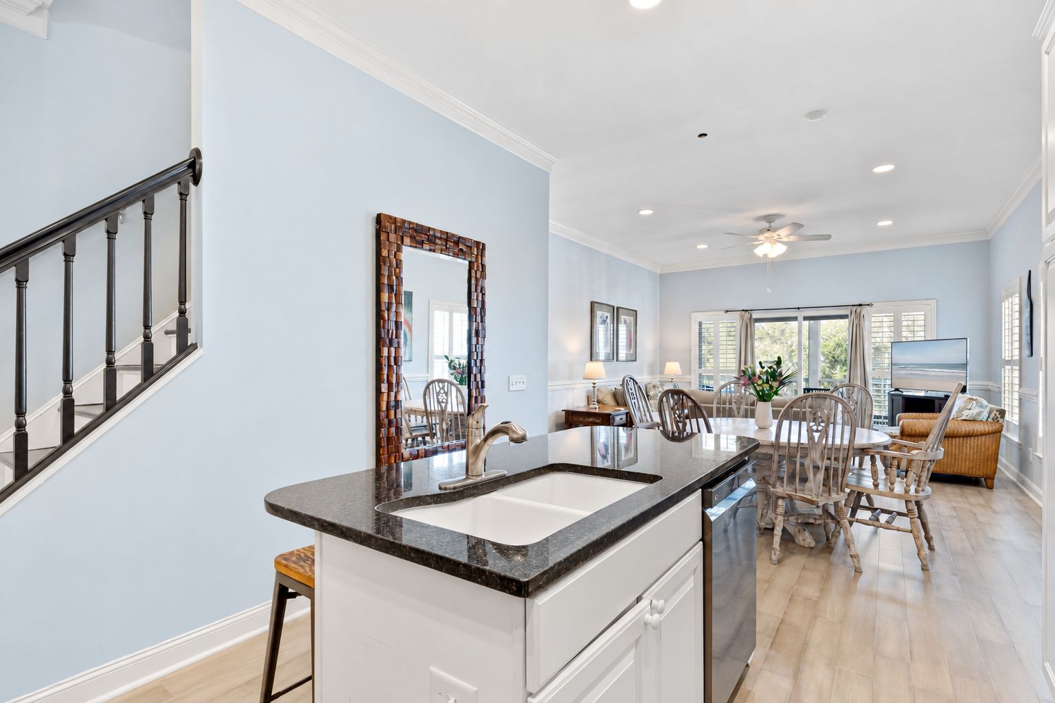 A modern kitchen with a granite countertop, a sink, and a dishwasher. There is a dining area with a table and chairs, and a living room with a sofa and a television.