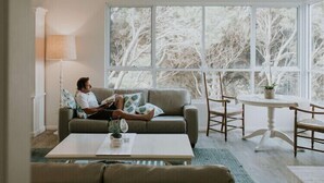 A cozy living room with a person reading, a sofa, a coffee table, and a large window with a view of trees. - Cliff Beach House, luxury Yallingup accommodation in secluded private estate (Yallingup)