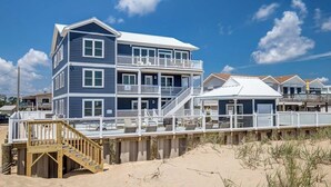 Una casa de playa de varios pisos con un balcón, una terraza de madera y una escalera que lleva a los niveles superiores.