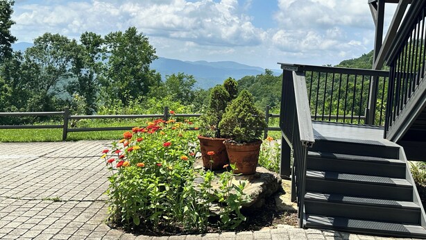 Un área exterior pavimentada con un banco de piedra, plantas en macetas y una escalera que conduce a un balcón con vistas a un paisaje montañoso.