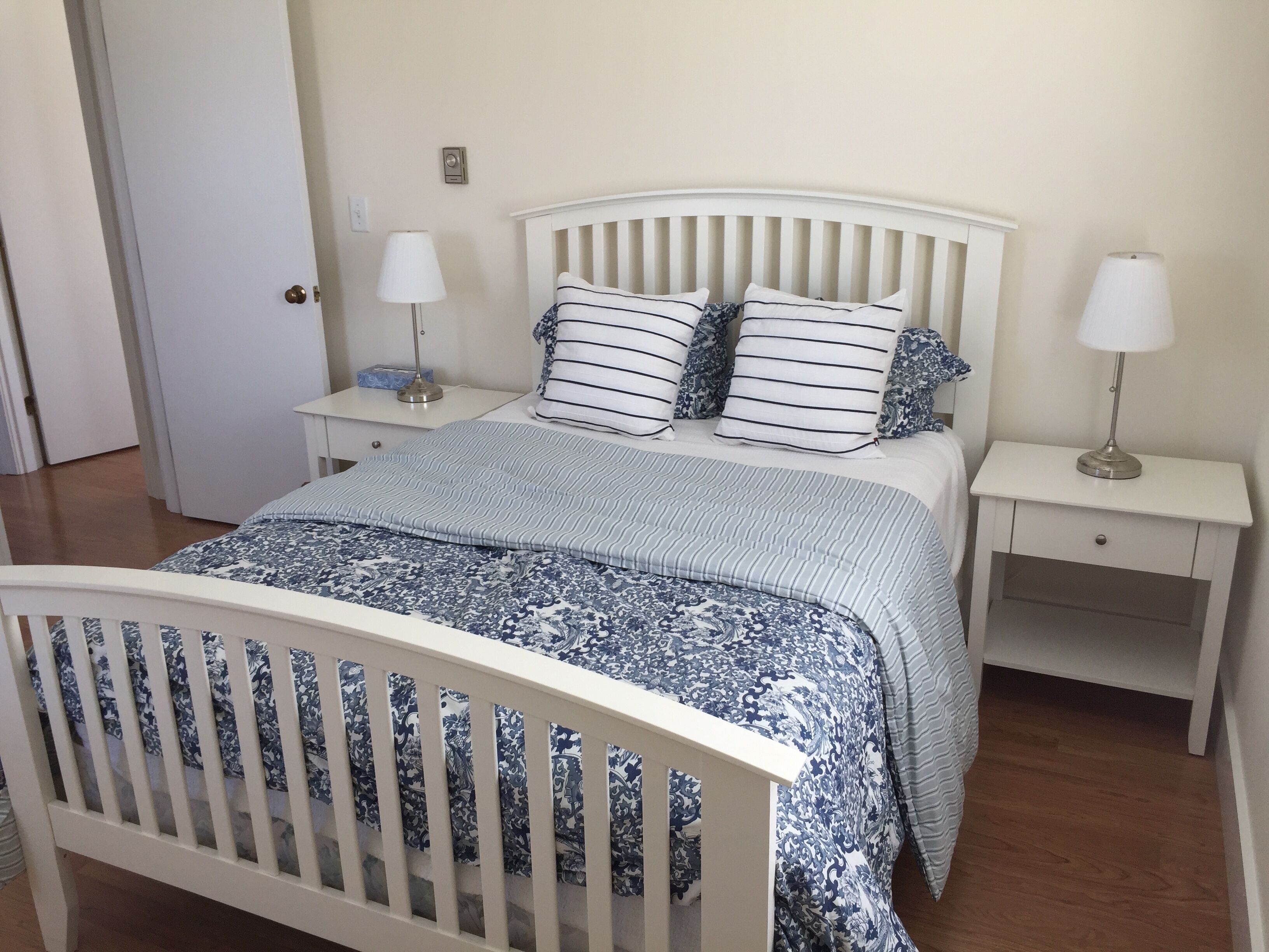 Une chambre avec un berceau blanc, un lit avec une literie à rayures, deux tables de chevet avec des lampes et une porte visible au fond.