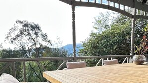 A wooden table with a woven chair, a metal roof, and a view of trees and mountains.