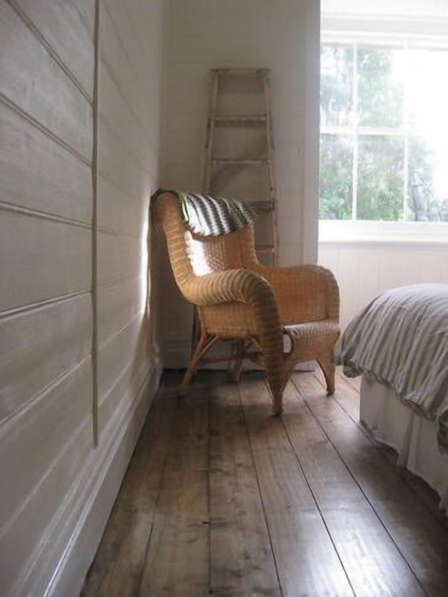 A corner room with a wooden floor, a wicker chair, a ladder, and a window with a view of trees.