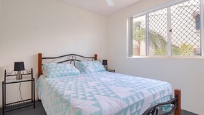 A single bed with a geometric patterned bedspread, two bedside tables with lamps, a window with a chain-link fence, and a view of palm trees outside.