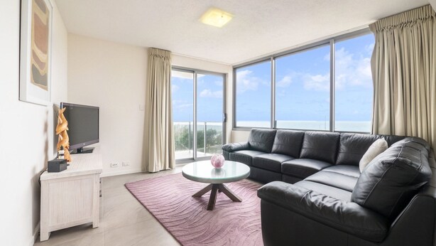 A modern living room with a black leather sofa, a glass coffee table, and a flat-screen TV on a white cabinet. Large windows offer a view of the sea. - Pacific Towers Unit 4, 19 Ormonde Tce Kings Beach (KINGS BEACH)