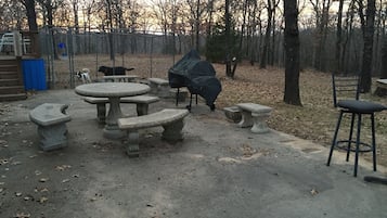 Ein Terrassenbereich im Freien mit Steintischen und -bänken, umgeben von Bäumen und einem Zaun.