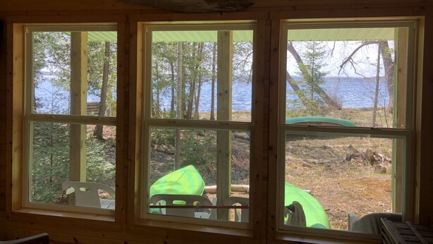 Una cabaña con tres ventanas amplias que ofrecen vistas a un bosque y un lago.