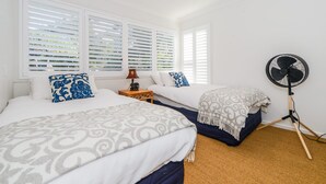 A bedroom with two beds, a standing fan, and a window with blinds.