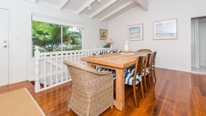A dining area with a wooden table and wicker chairs, white walls, and a view of greenery through a large window.