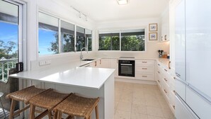 A modern kitchen with white cabinetry, a central island with stools, a dishwasher, and a large window offering a view of trees.