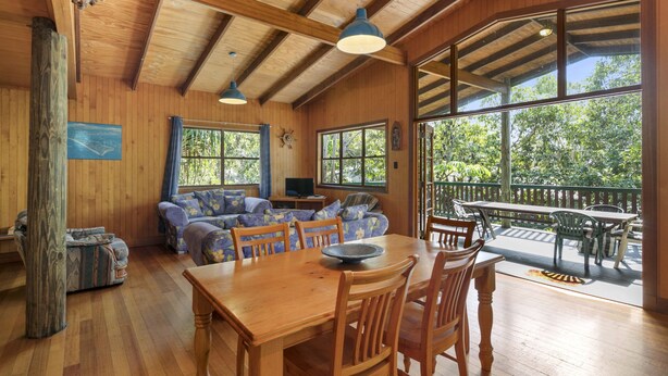 A wooden interior with a dining table and chairs, a sofa, and large windows offering a view of greenery.