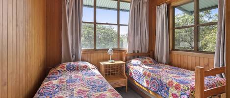 A cozy cabin room with two wooden beds, a small table with a lamp, and large windows with curtains.