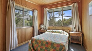 A cozy bedroom with wooden walls, a quilted bedspread, two windows with curtains, and a ceiling fan.